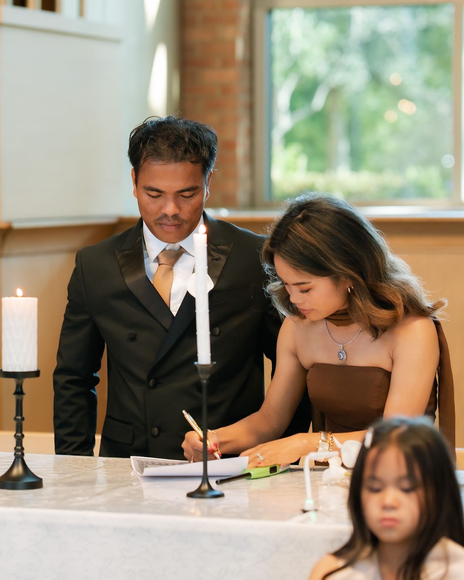 Couple signing wedding documents at a white table with candles, woman in brown dress and man in black suit.