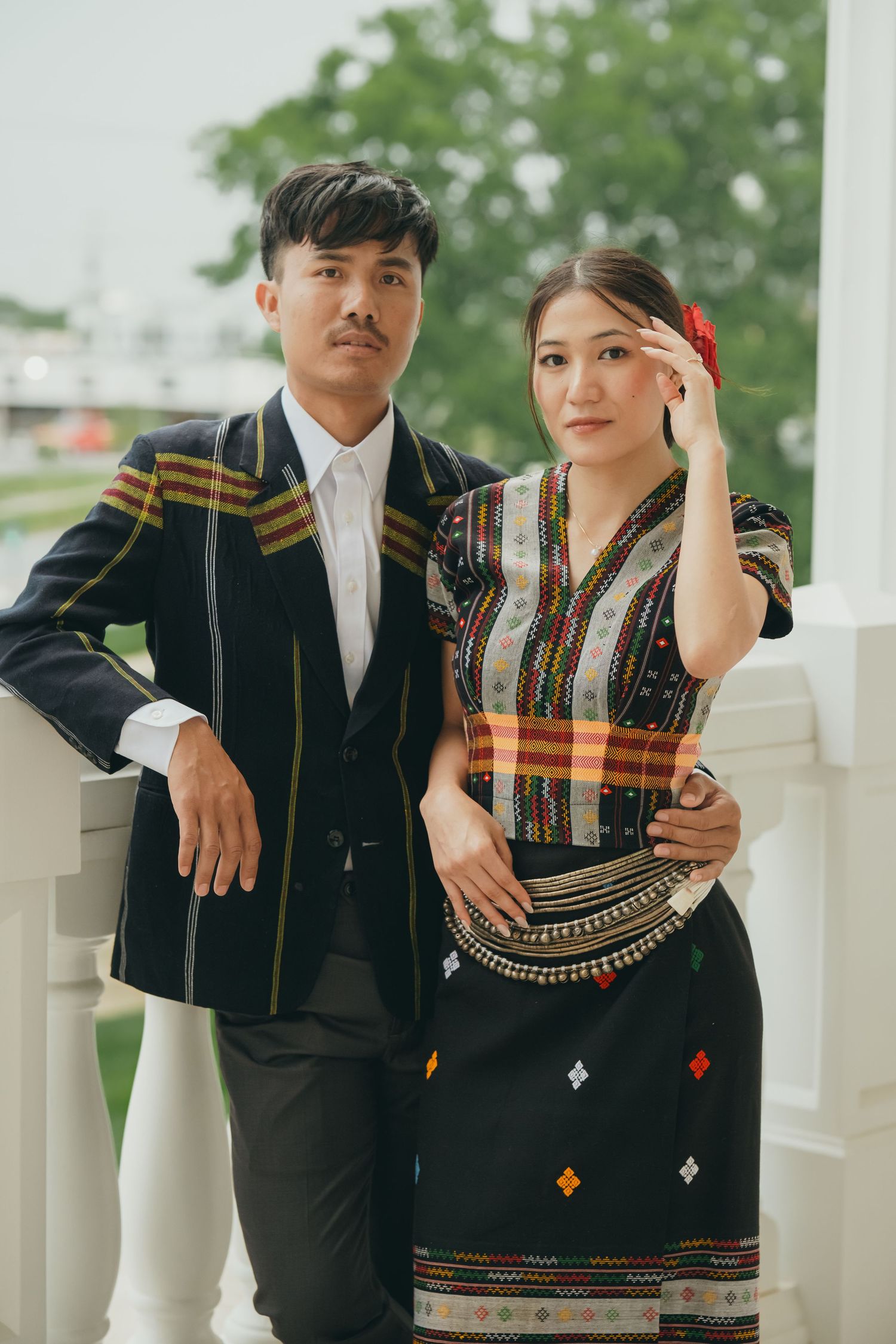 Couple in traditional Hmong attire poses together near white columns outdoors.