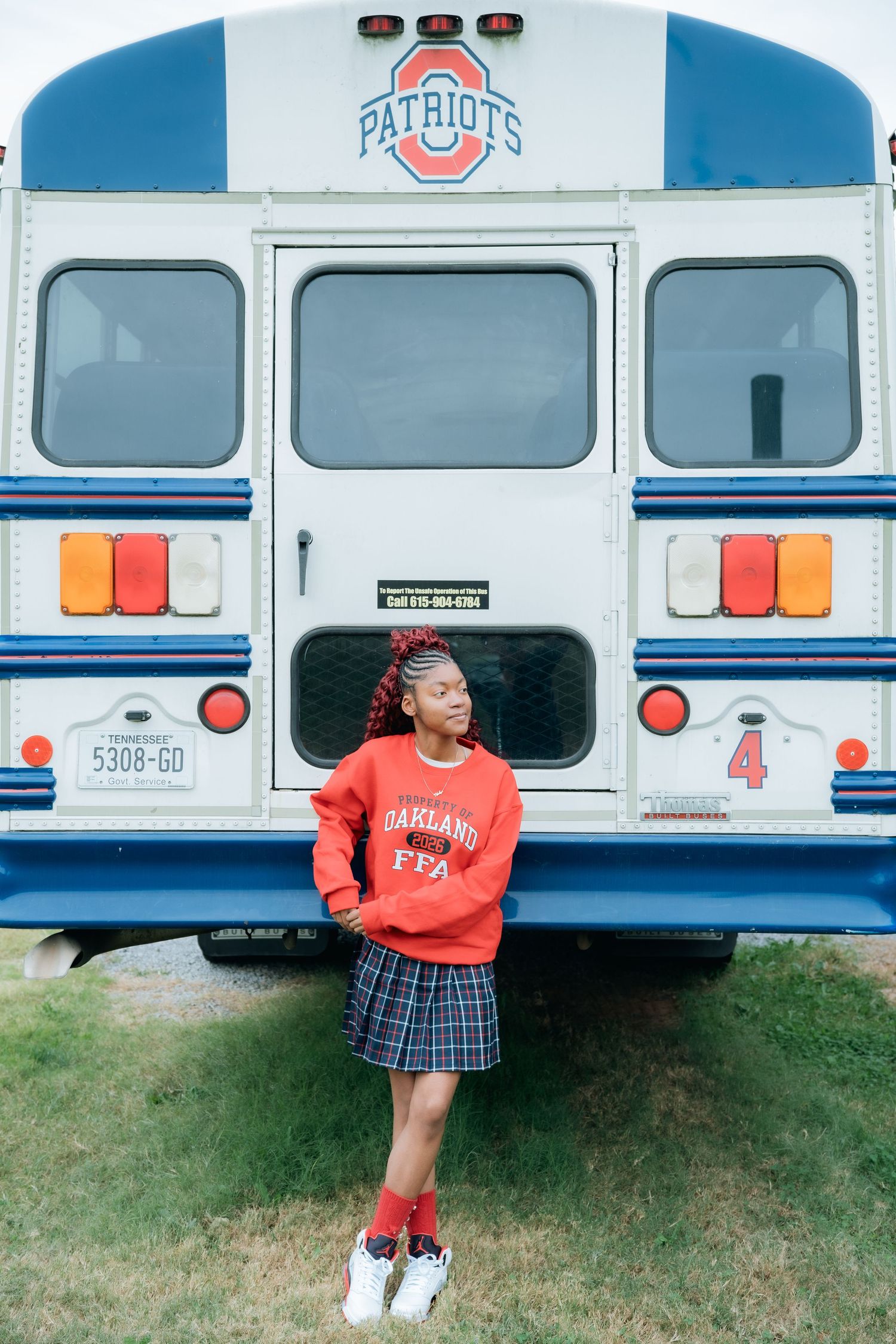 Student in school uniform poses beside white and blue Patriots school bus.