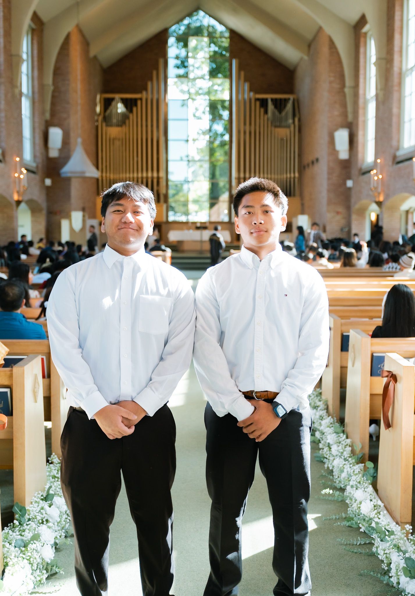 Two young men in white shirts and black pants stand in a church aisle decorated with greenery during a wedding ceremony.