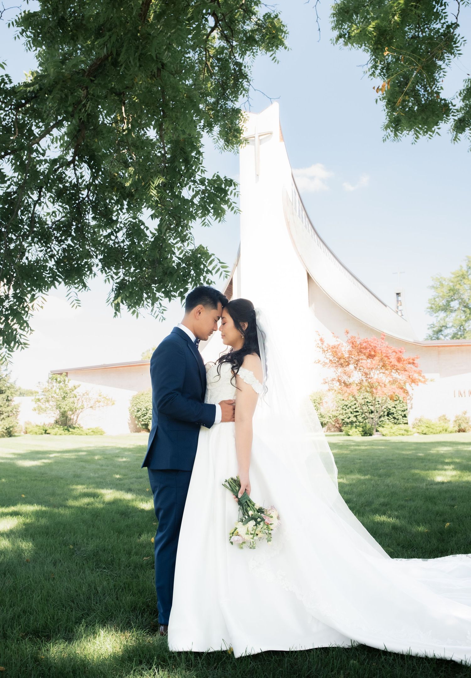 Romantic wedding portrait in front of modern white church architecture with trees framing the scene.