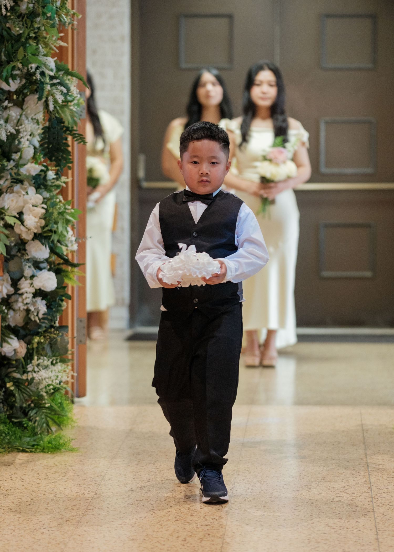 Young ring bearer in formal vest and pants walks down aisle carrying wedding rings.