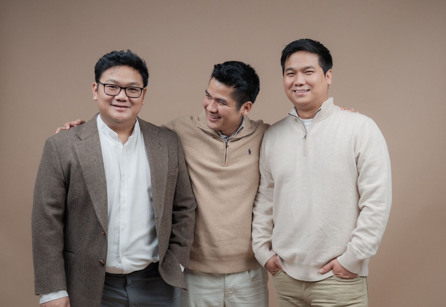 Three people in coordinated neutral-colored outfits share a candid moment against a brown backdrop.