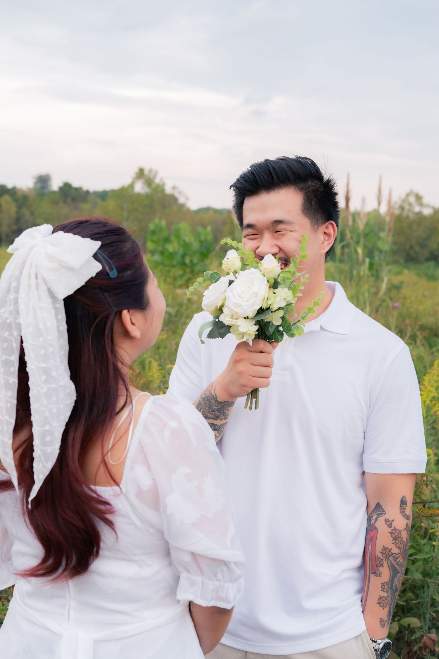 A couple in white outfits poses playfully with flowers in a natural outdoor setting.