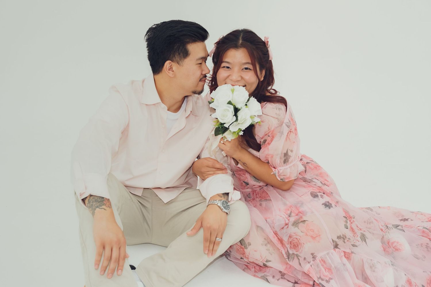 Couple embraces while wearing pink floral dress and light colored shirt in studio.