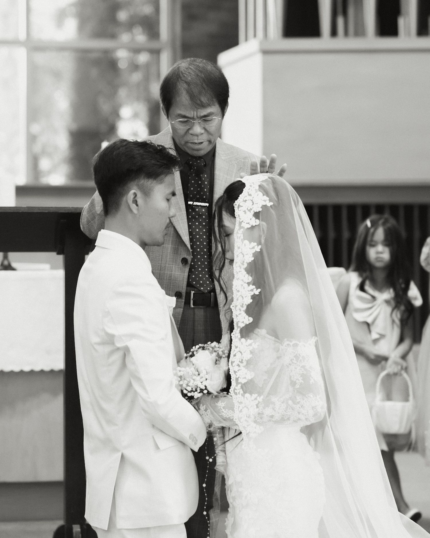 Black and white wedding ceremony photo of couple exchanging vows before officiant, bride in lace veil holding bouquet.