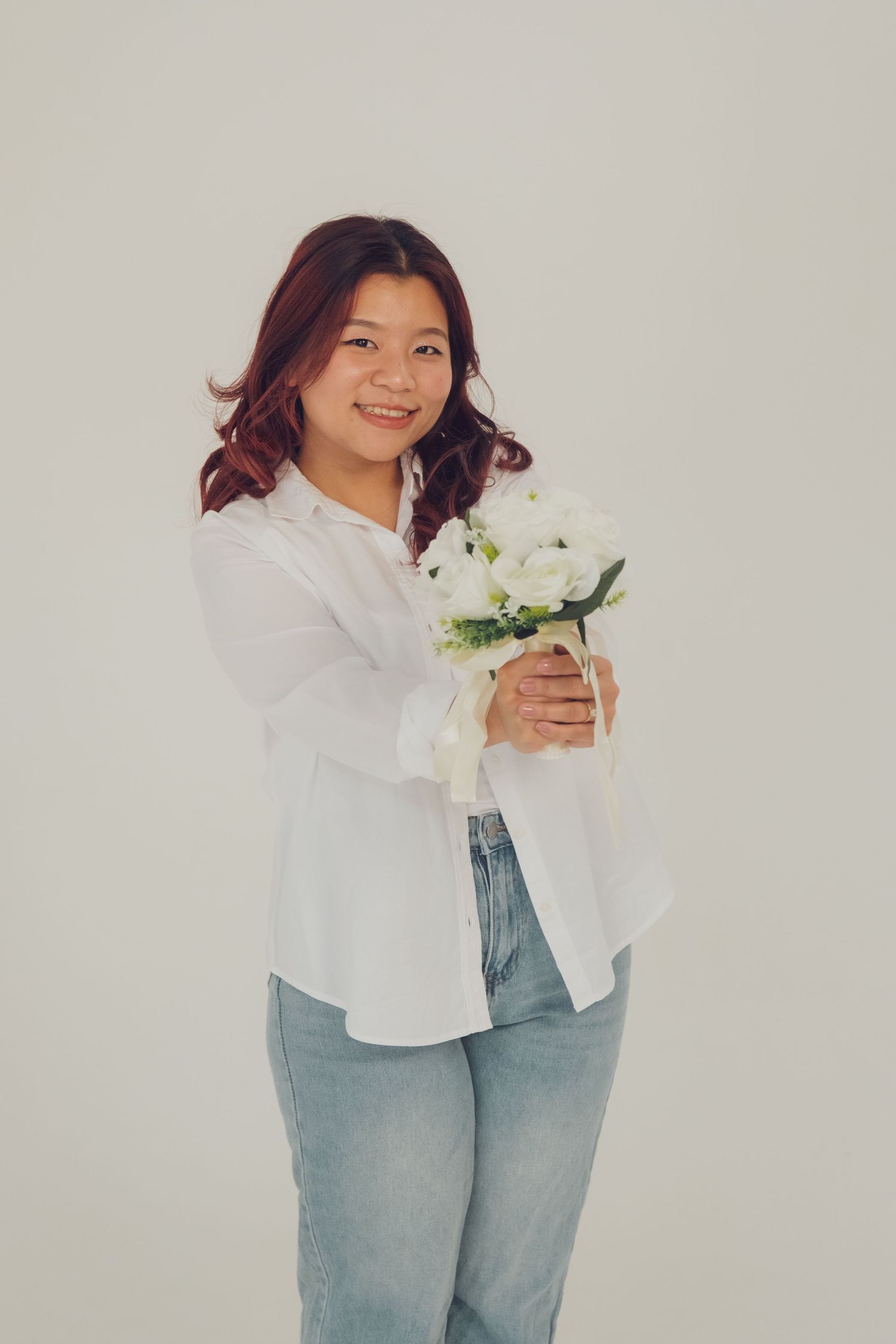 Woman in casual white shirt and jeans poses with white flowers against light grey wall.
