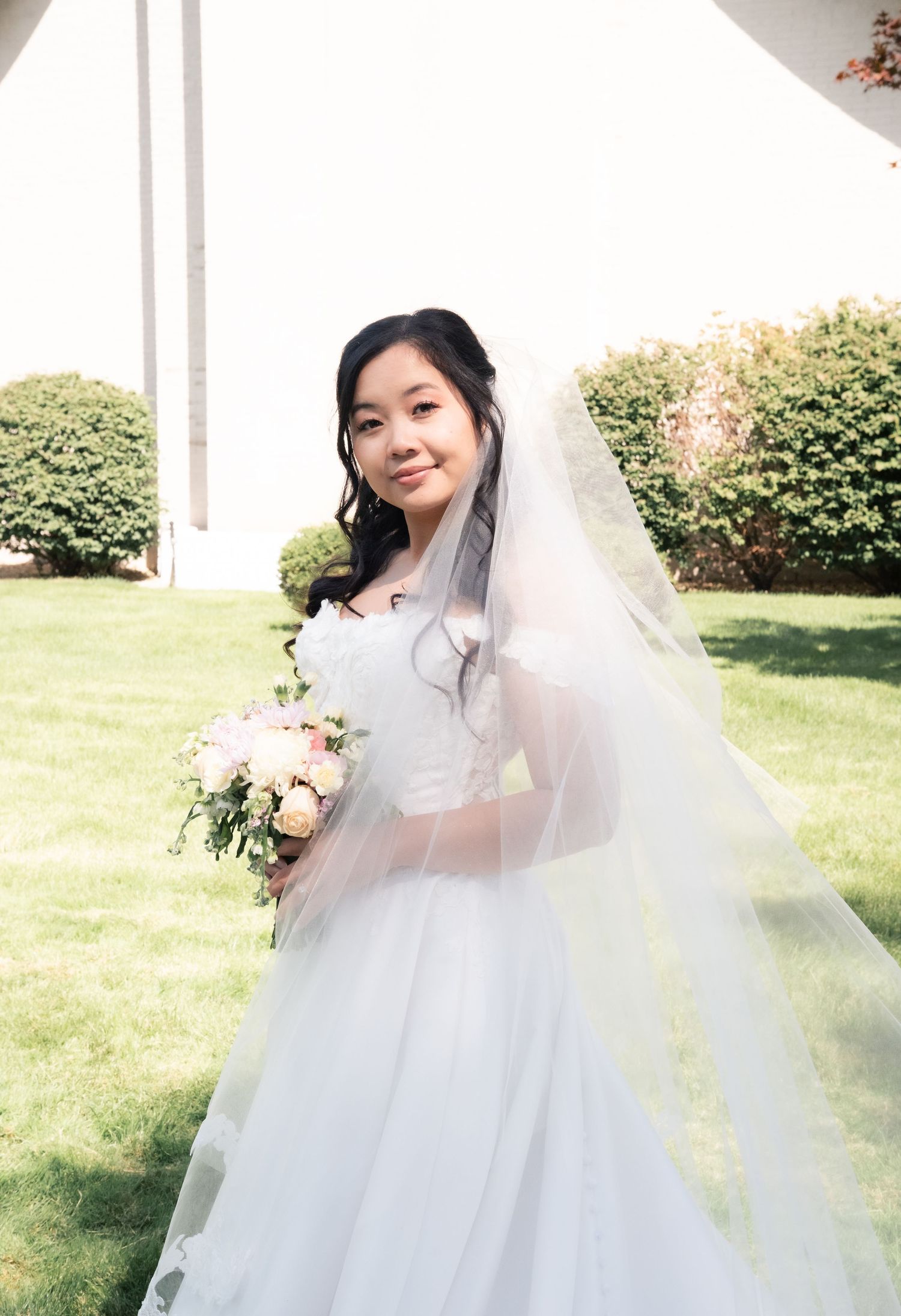 Bride in white wedding dress and veil poses with bouquet on sunny lawn with greenery in background.