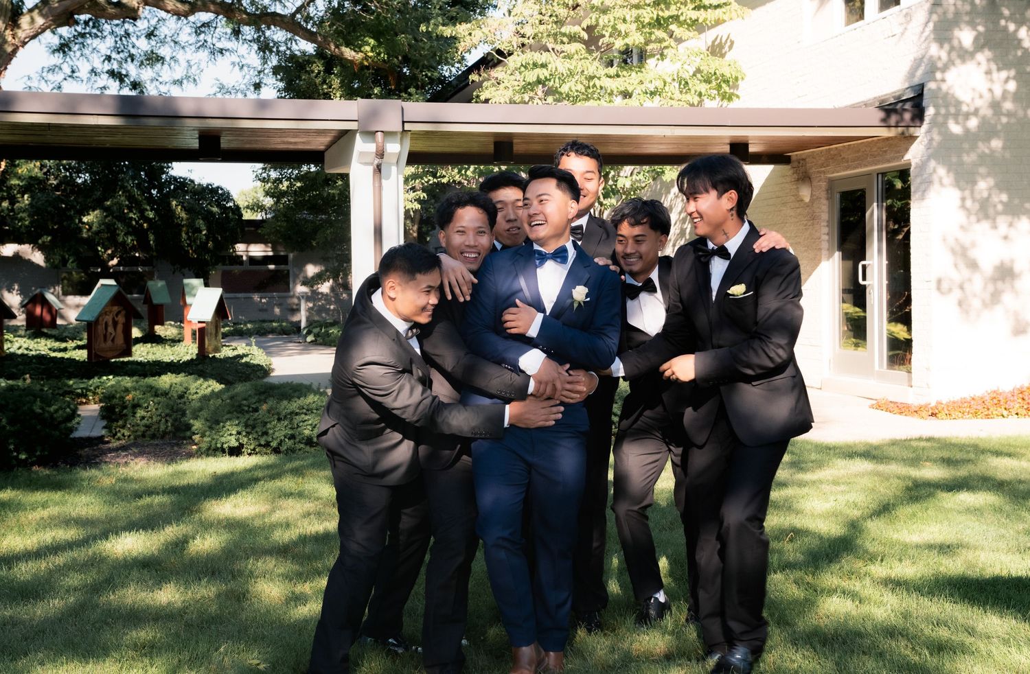 Wedding party members gather casually on grass near a building.