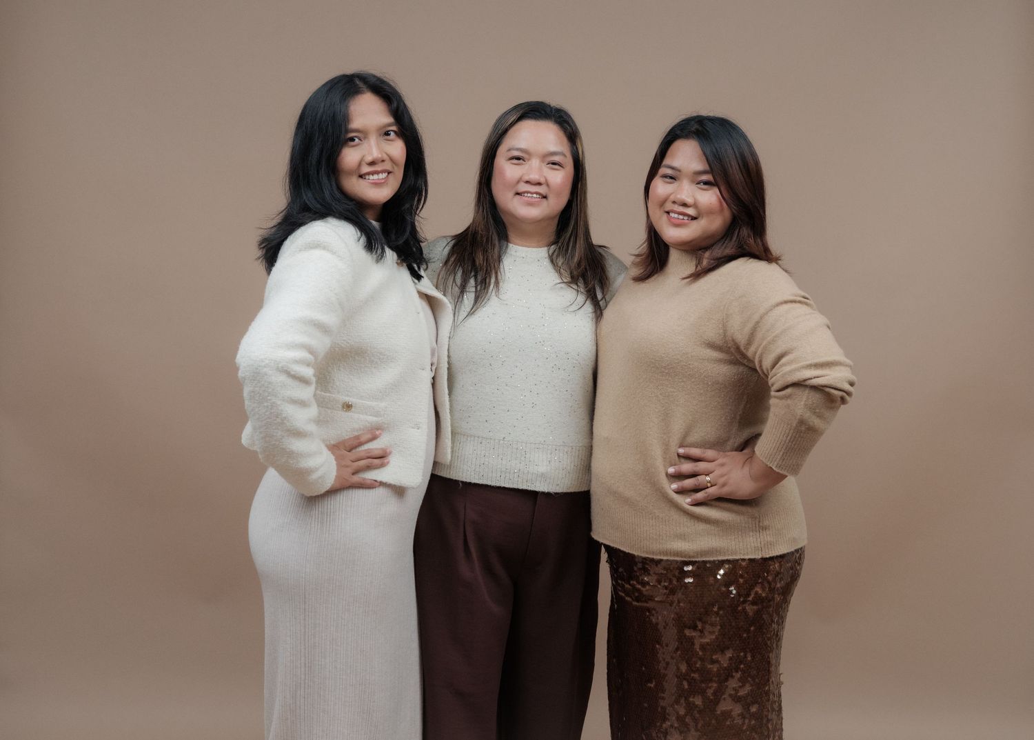 Three people in cozy sweaters pose together against a brown backdrop for a casual portrait.