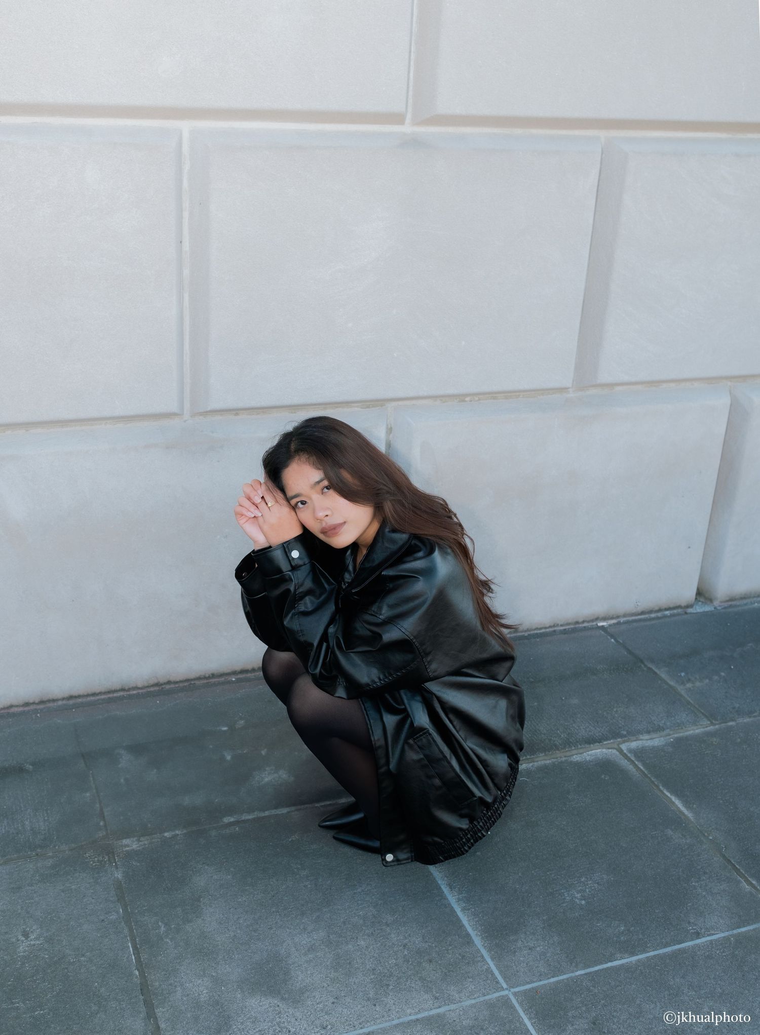 A person in a black leather jacket crouches against a light gray wall on dark stone flooring.