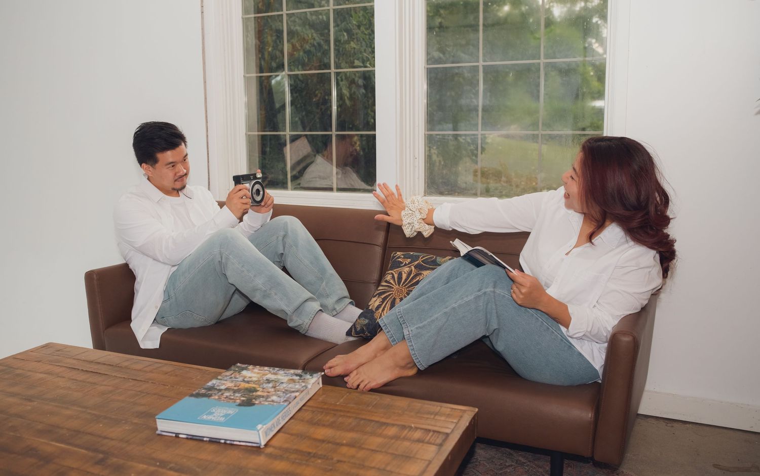 Couple in white shirts and jeans plays around on brown leather couch near window.