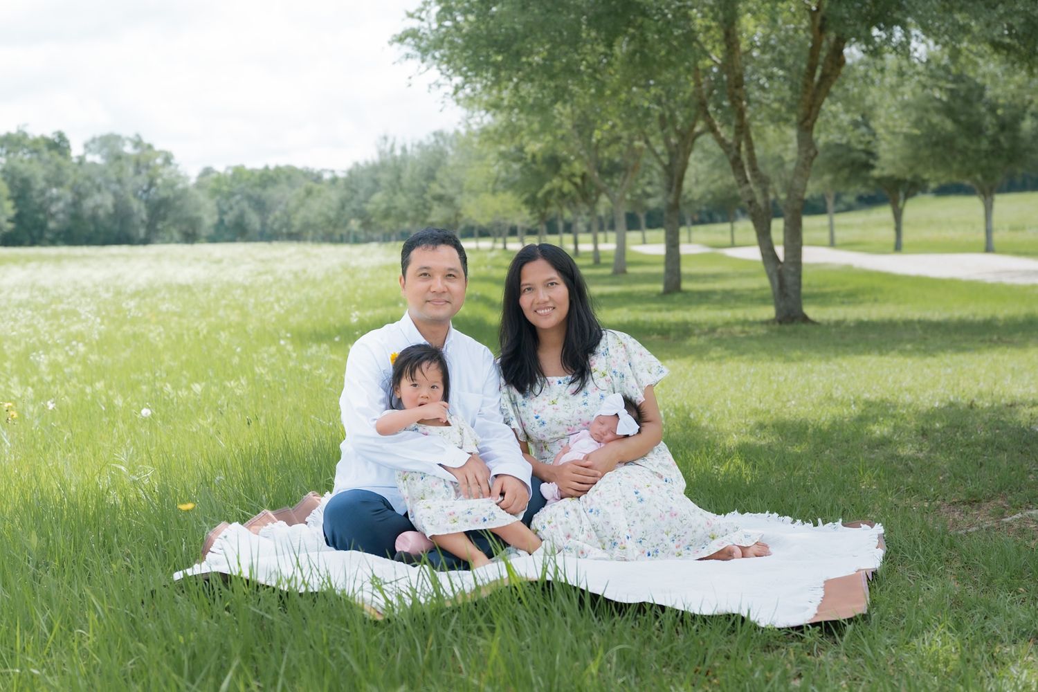 Happy family of four sitting on a blanket in a green park, parents holding young children on a sunny day.