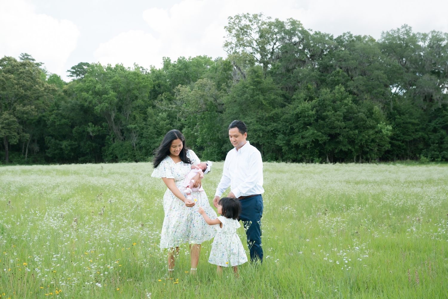 Pregnant woman in white dress with husband and young daughter in a lush green meadow during a family maternity photo session.