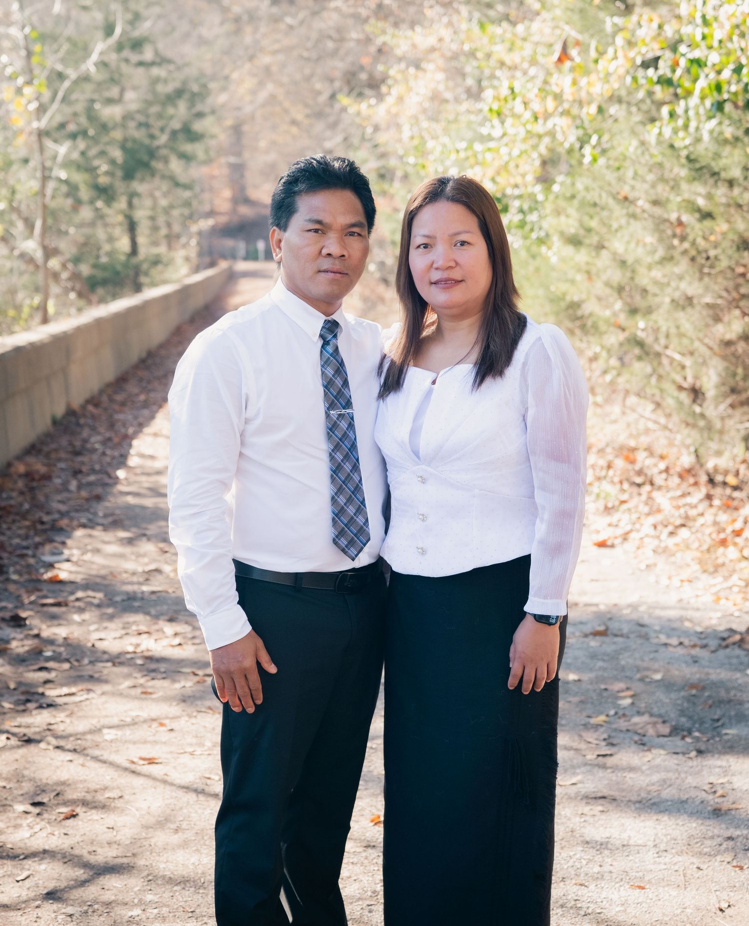 A couple in matching white shirts and dark formal wear poses together on a sunlit outdoor path.