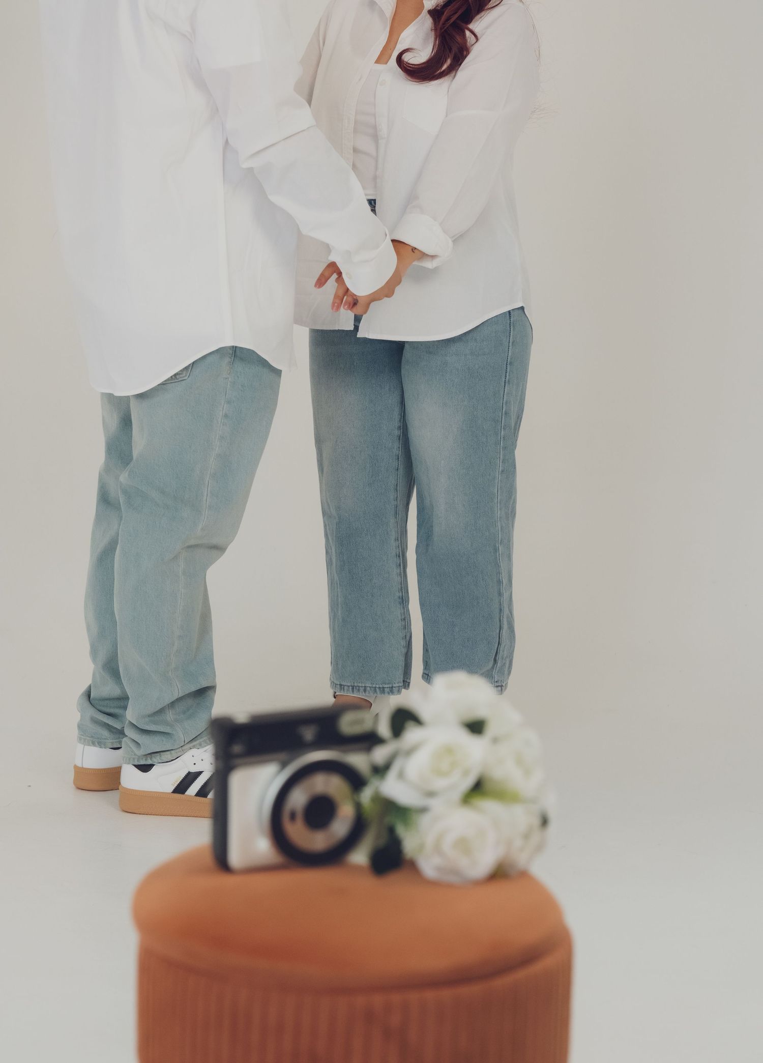 A couple in matching white shirts and jeans stands together against a white background with flowers.