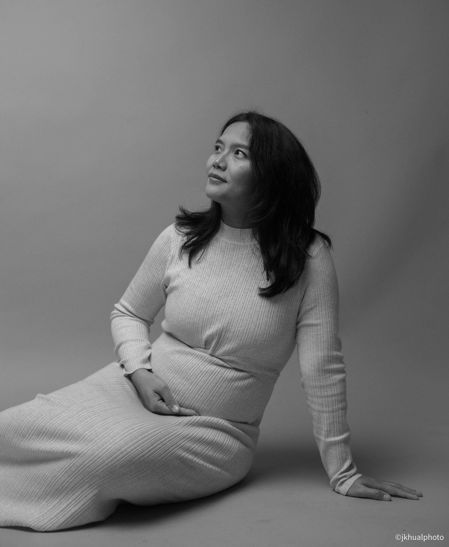 Black and white portrait of pregnant woman in ribbed sweater dress sitting on studio floor.