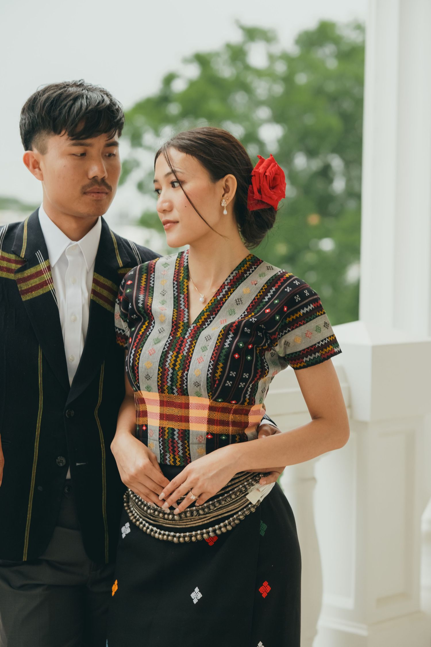 A couple in traditional Asian formal wear poses together with the woman wearing a red flower.