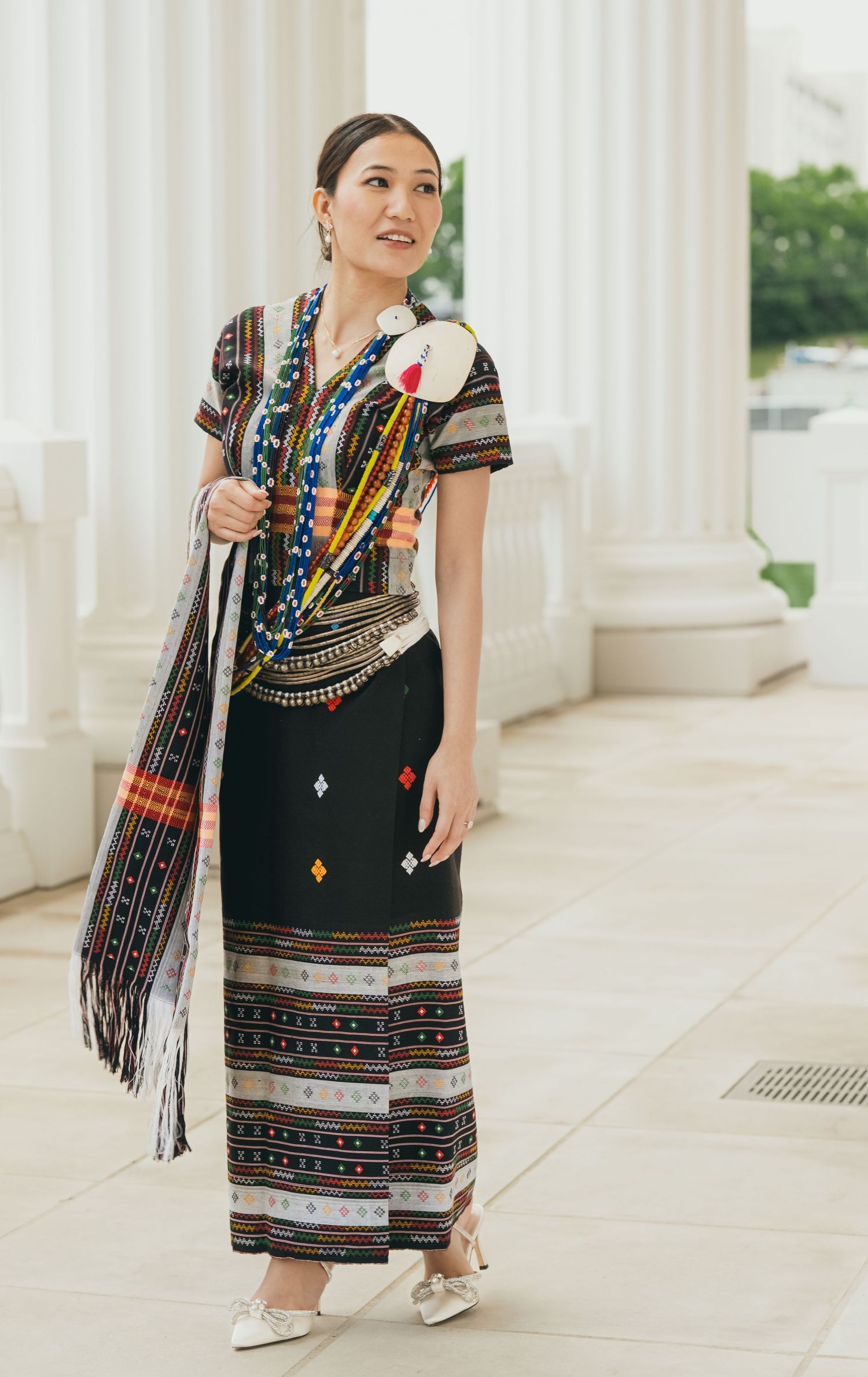 Colorful traditional dress with intricate patterns and beadwork shown against white pillared background.