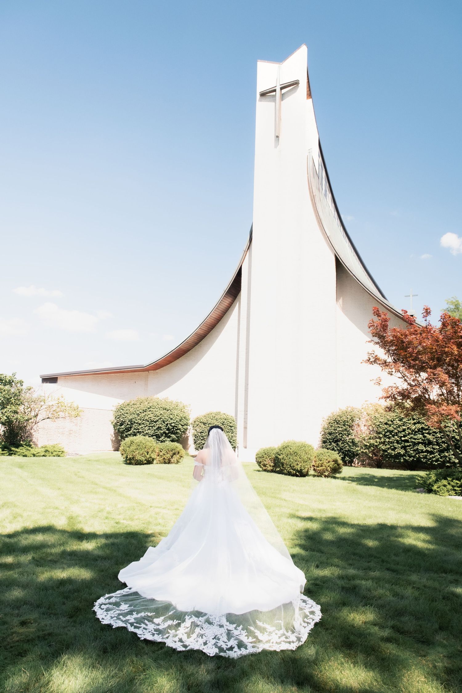 White lace wedding dress train spreads across grass near modern church building.