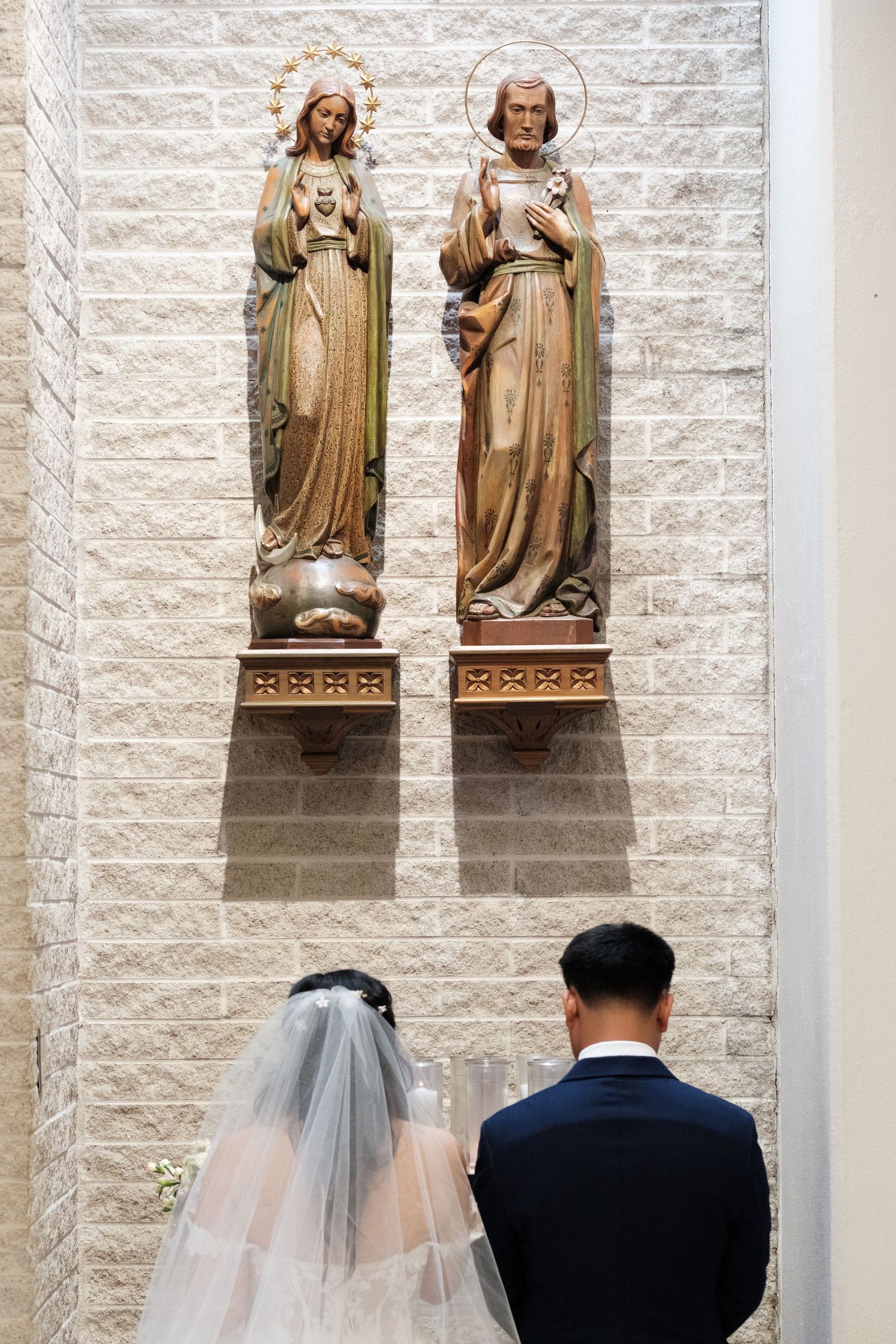 A couple prays beneath two religious statues mounted on a white brick wall.