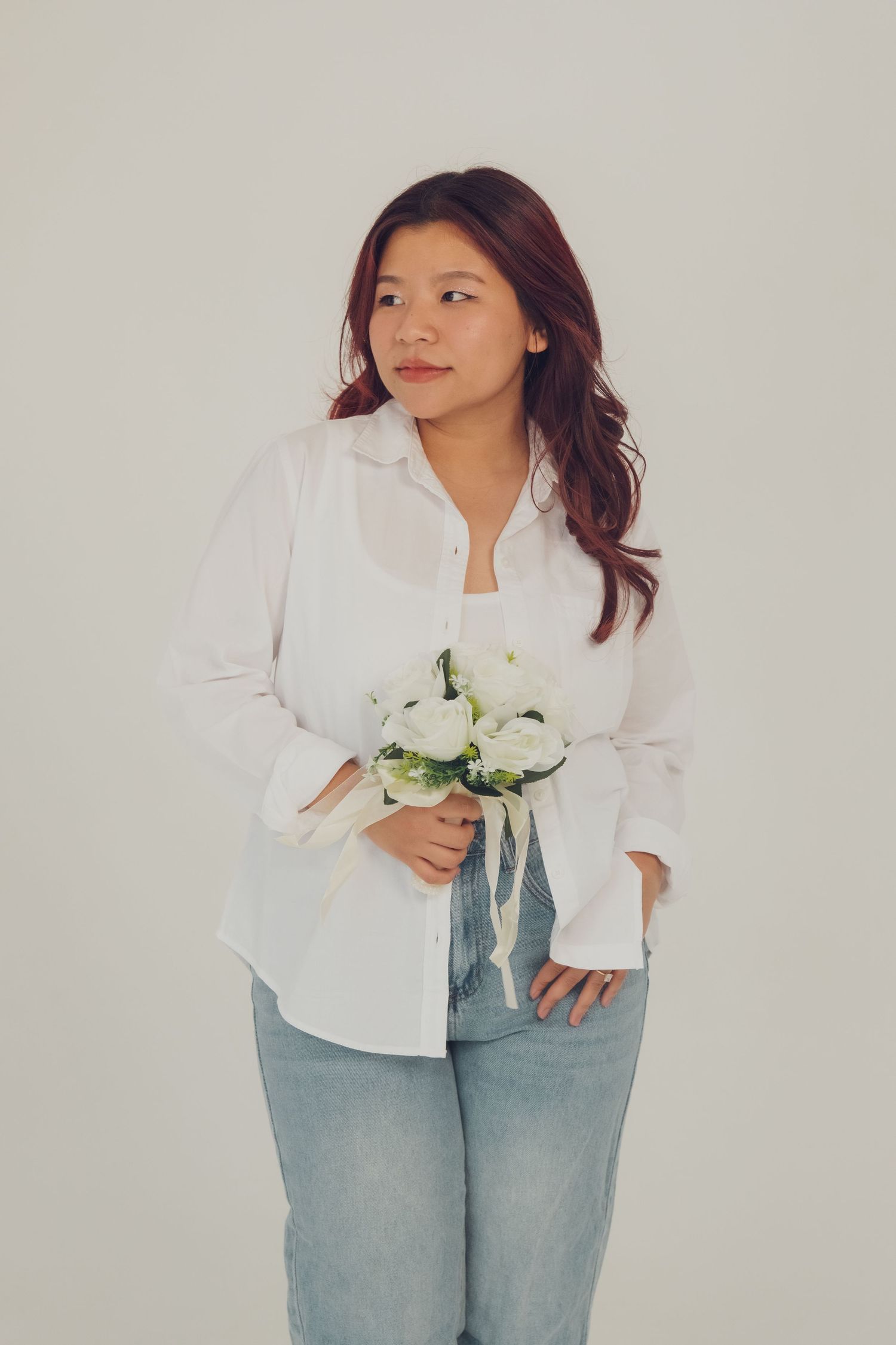 Woman in white blouse and jeans holds white flowers while posing against neutral background.