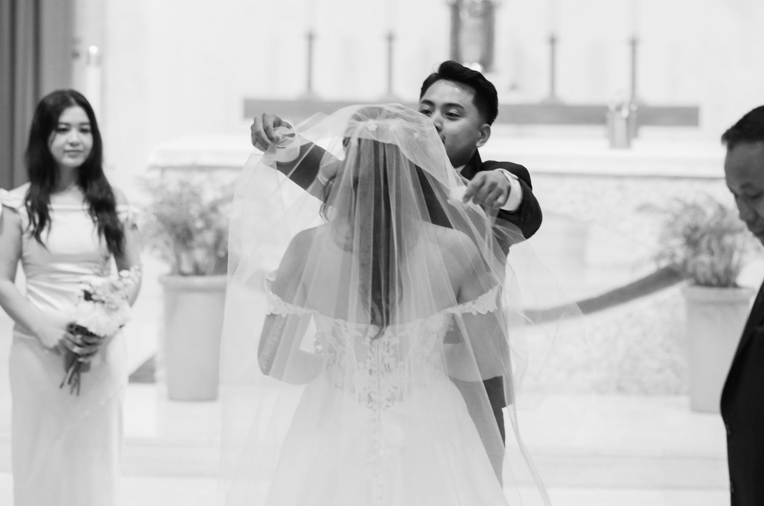 Black and white candid moment of veil adjustment during wedding ceremony.