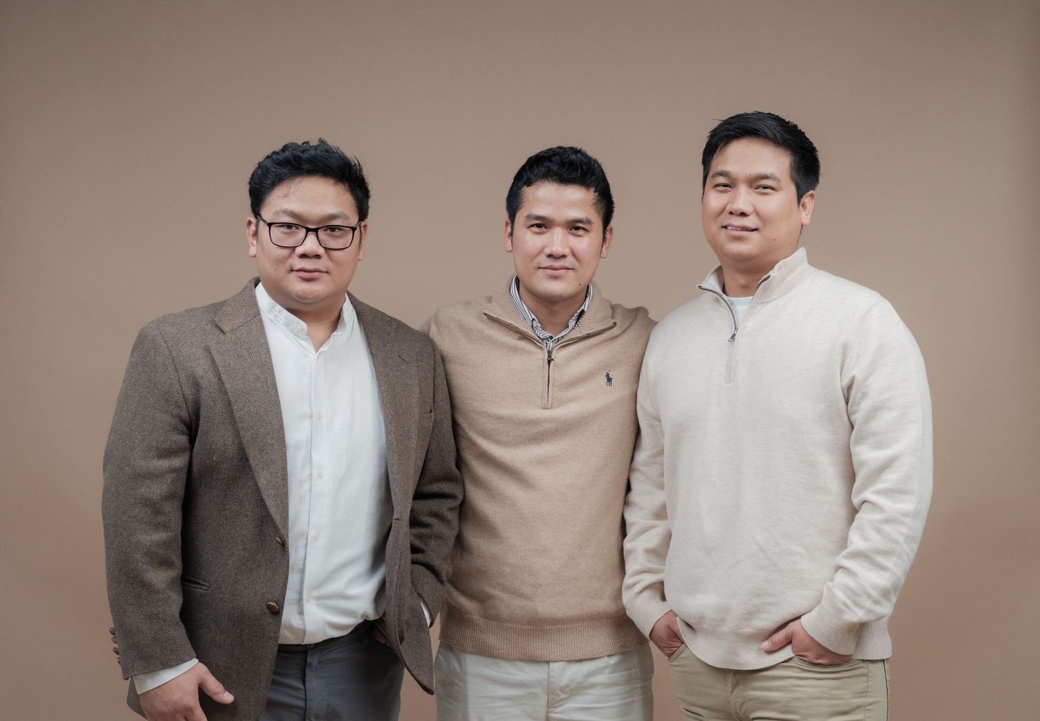 Three people in neutral-toned clothing pose professionally against a brown studio backdrop.
