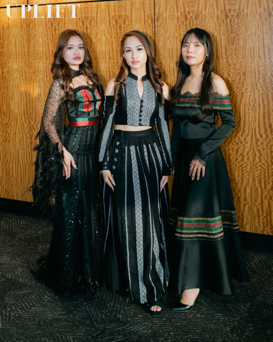 Three friends in coordinated black and patterned dresses pose together against a wooden wall.