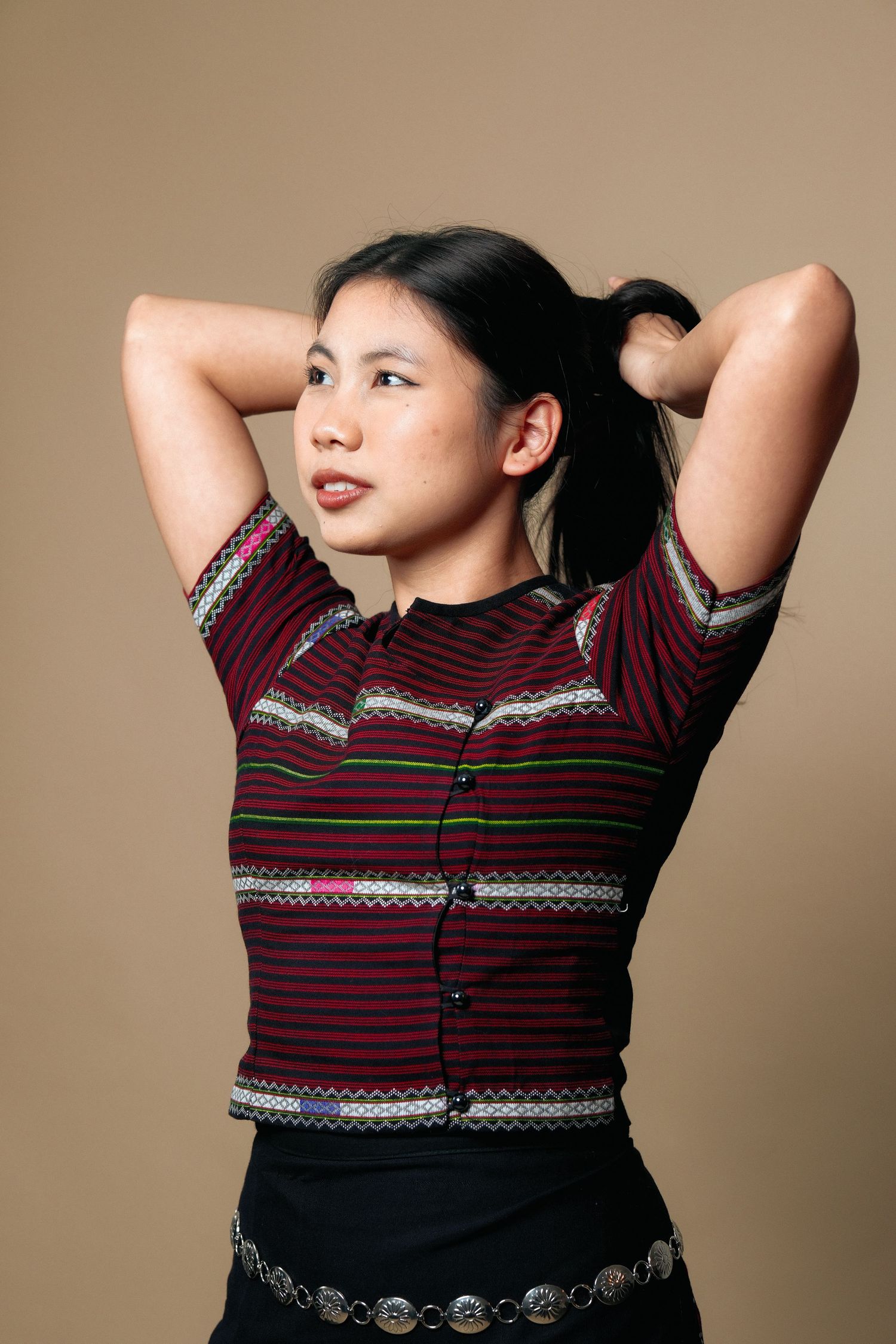 Model wearing burgundy striped top with decorative trim and metallic belt against beige background.
