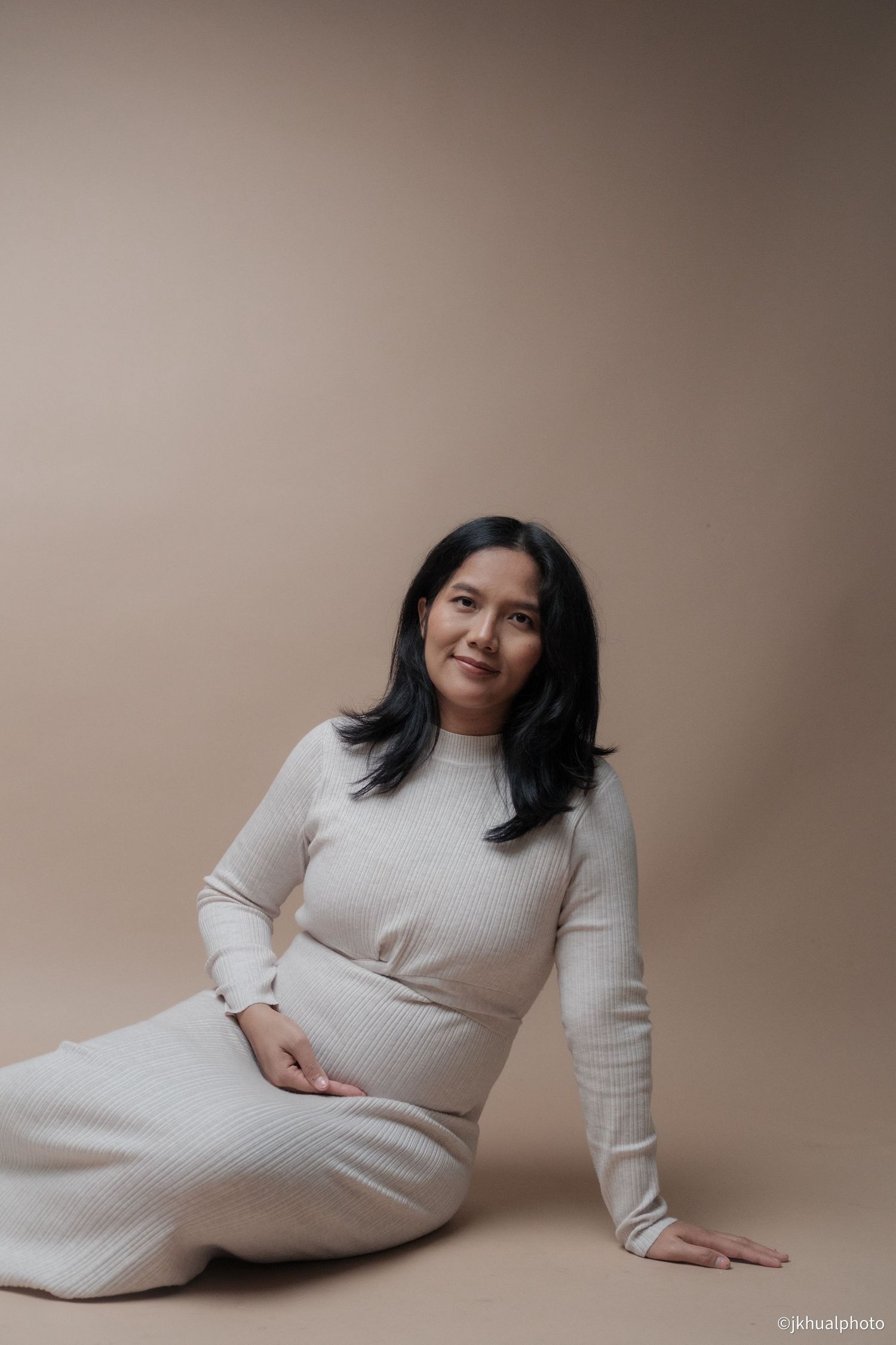 Maternity photo series showing a woman in a white knit dress posing against a neutral beige backdrop.