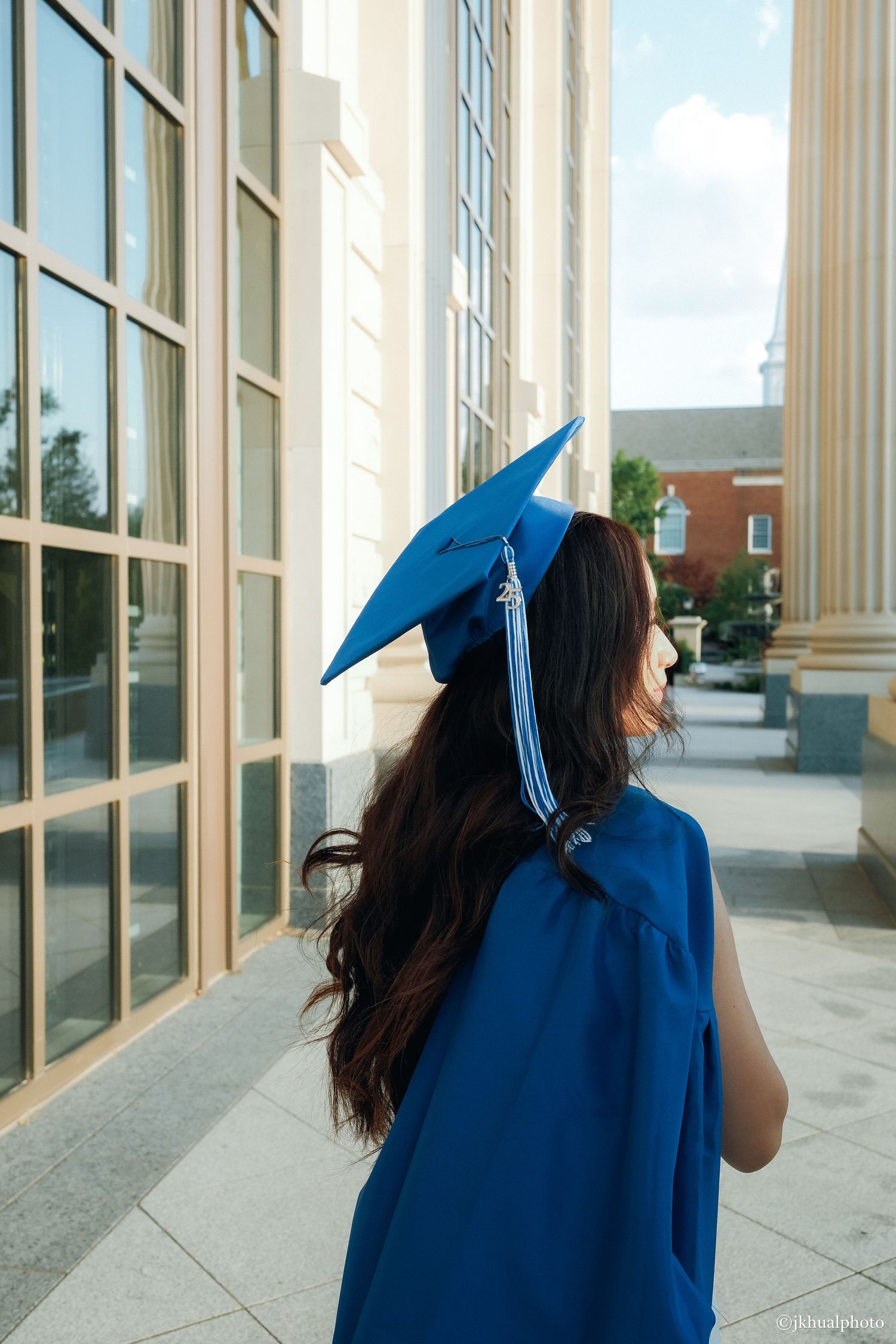 Graduate in blue cap and gown stands beside white pillars and windows.