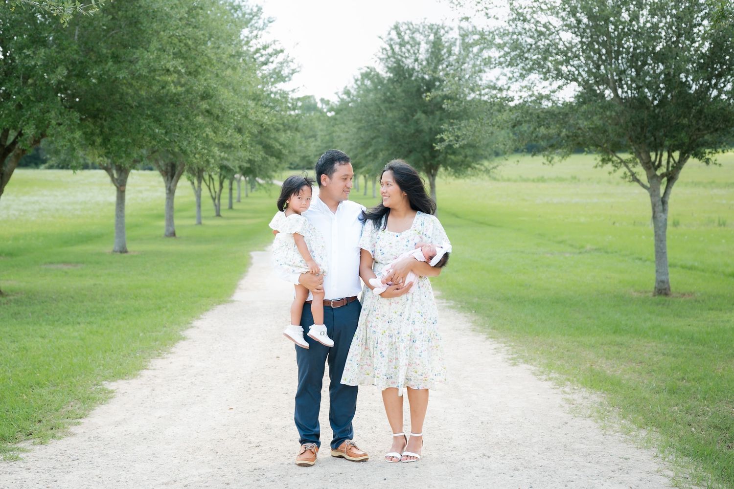 Family of three walks on a gravel path surrounded by lush green trees in a park.
