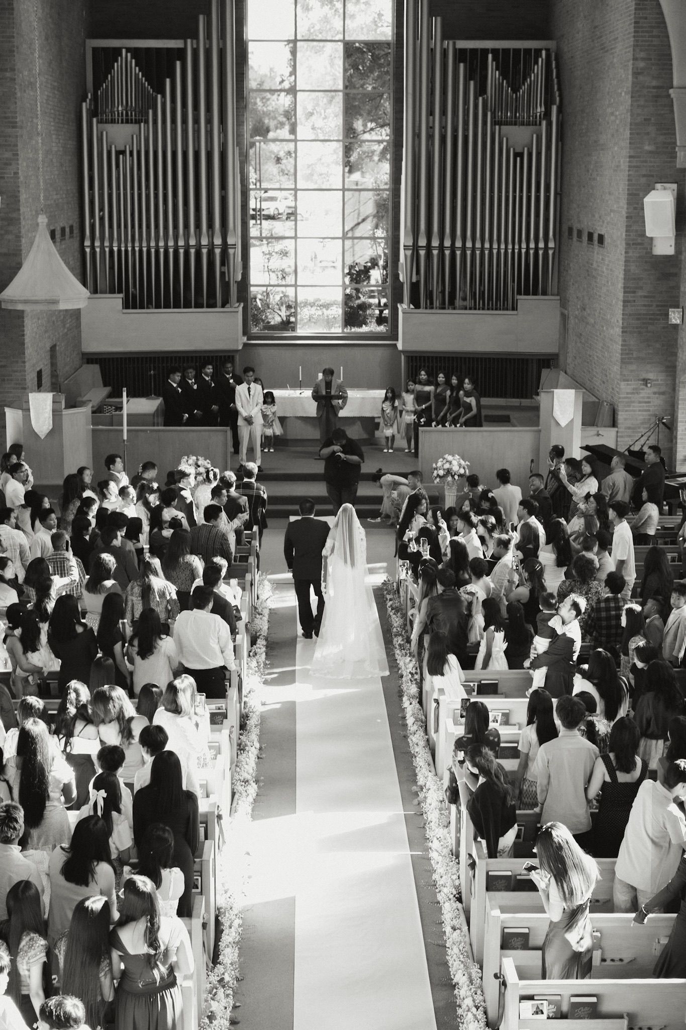 Black and white photo of bride and groom walking down church aisle surrounded by standing wedding guests.