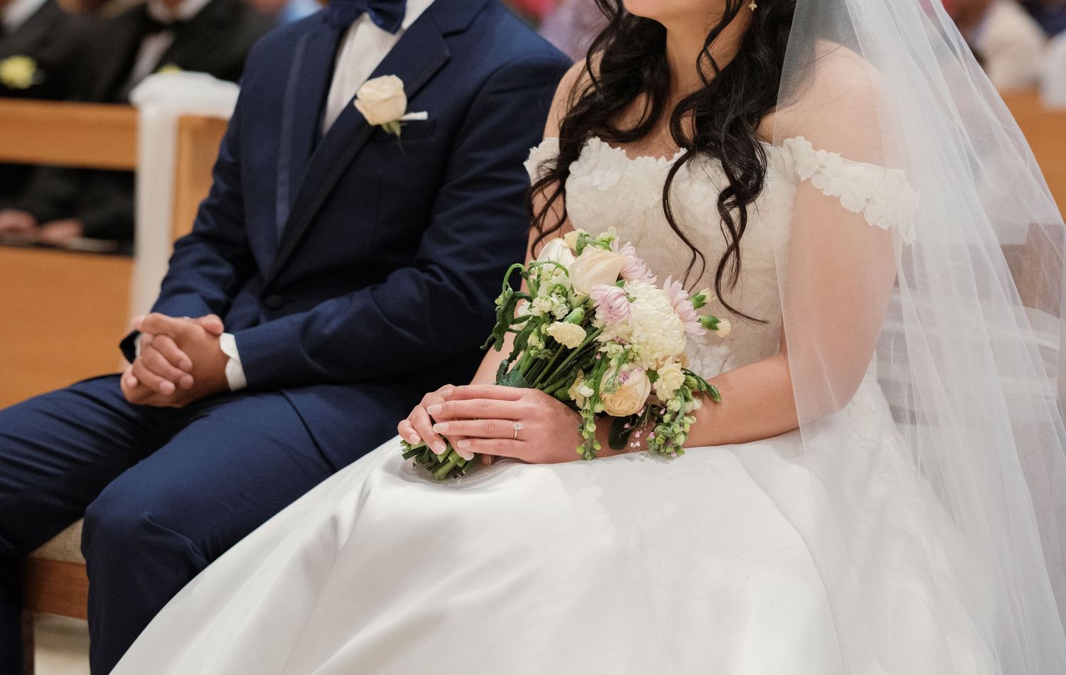 A bride in a white dress and veil sits during a traditional Catholic wedding ceremony.