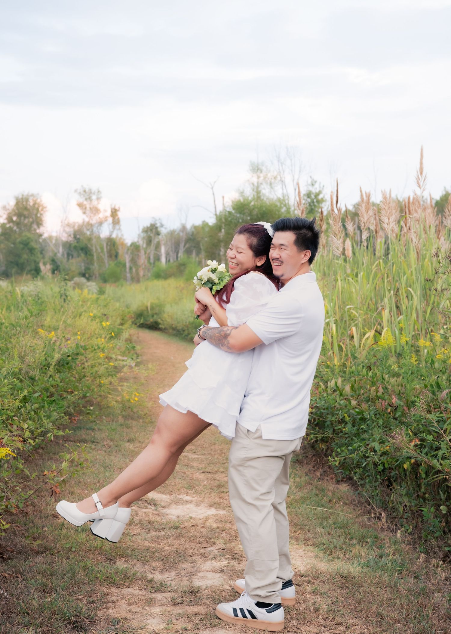 A couple in matching white outfits performs a playful lift pose in a natural setting.