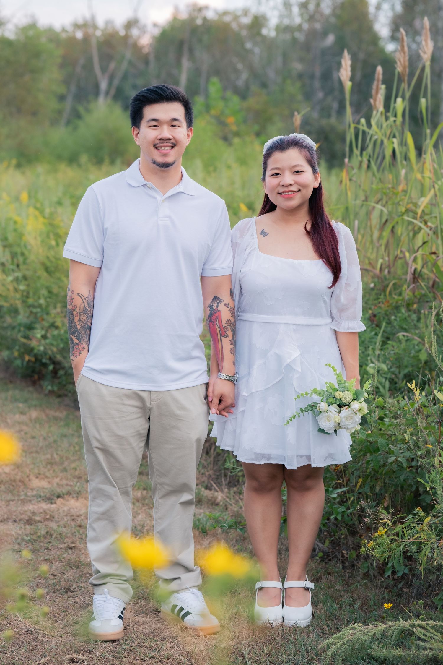 A couple in white outfits poses together on a nature trail with wildflowers and tall grass.