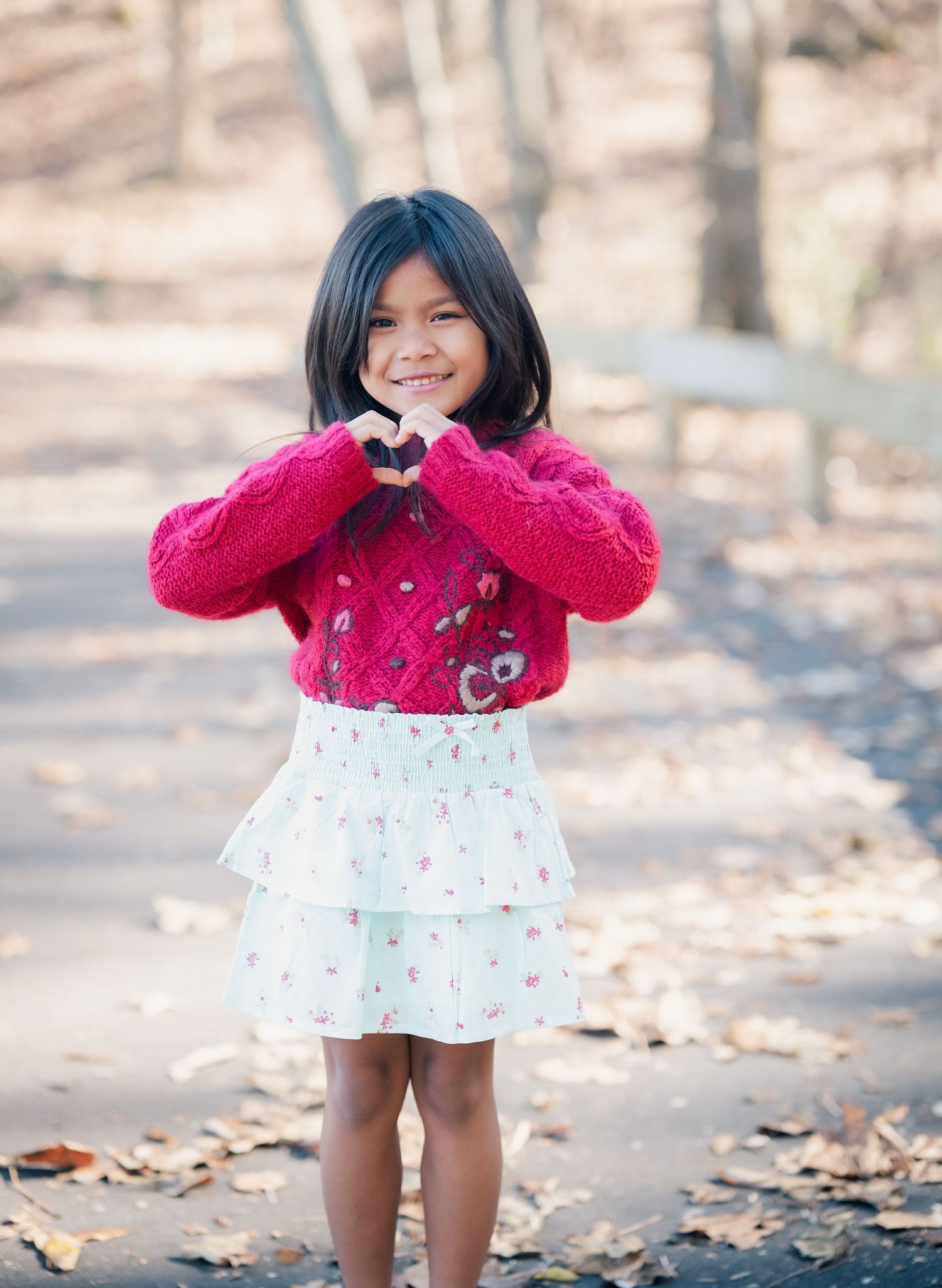 Young child wearing a pink sweater and white floral skirt makes heart shape with hands on autumn path.