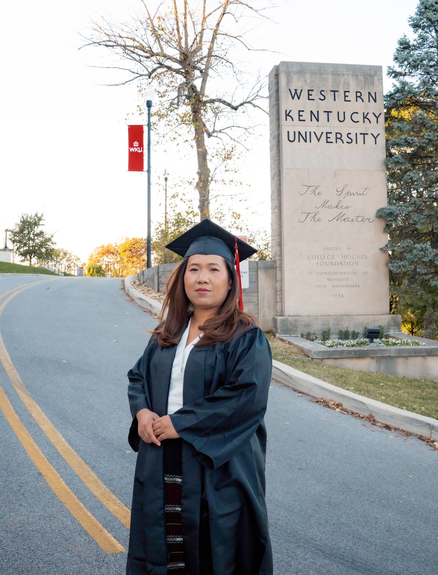 Graduate in cap and gown posing on campus road with university signage.