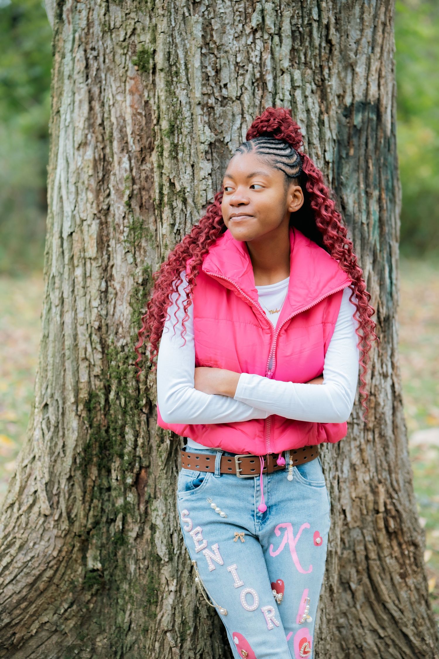 Person wearing pink puffy vest and decorated jeans posing against large tree trunk.