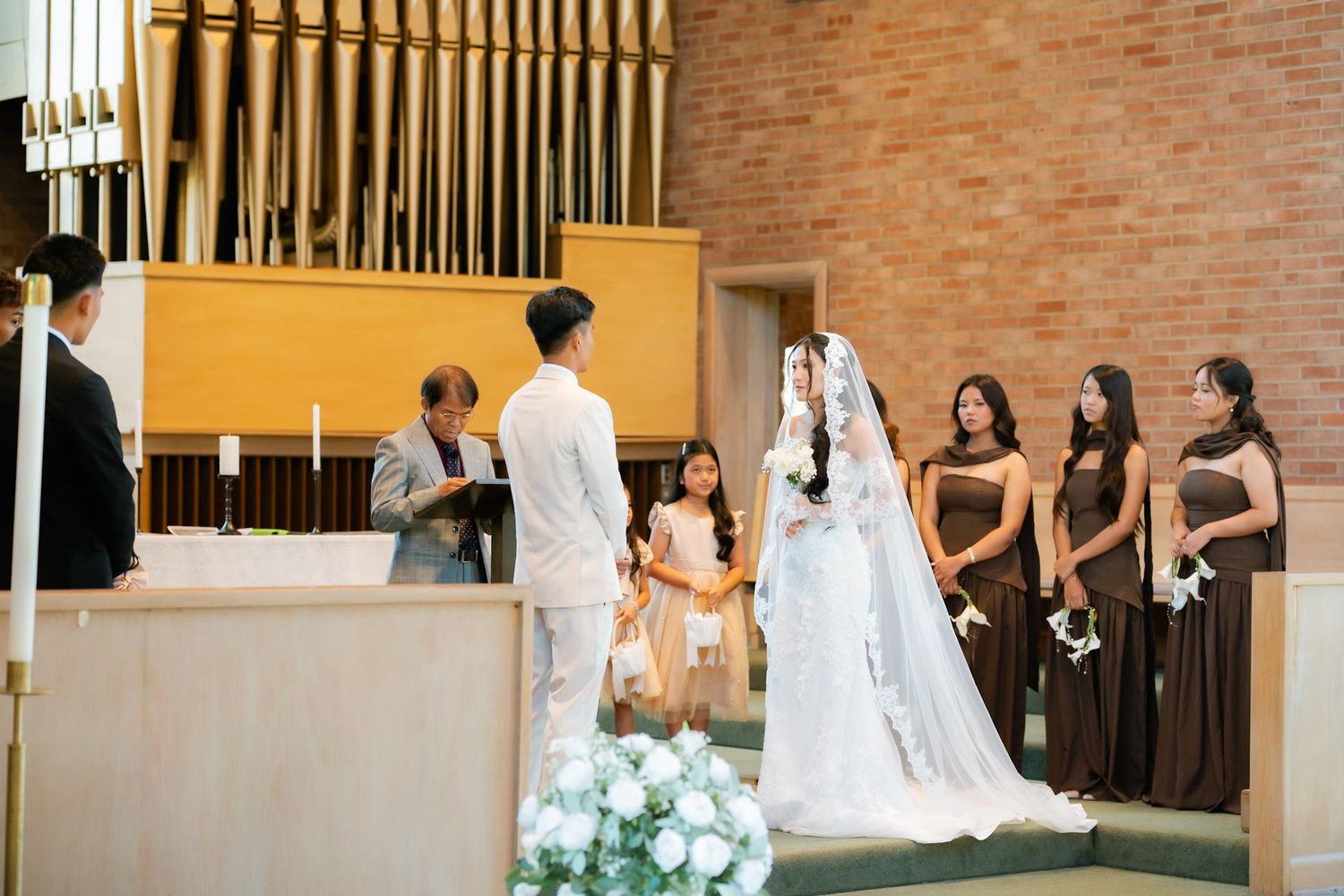 Couple exchanging vows at church altar with bridesmaids in brown dresses during elegant wedding ceremony.