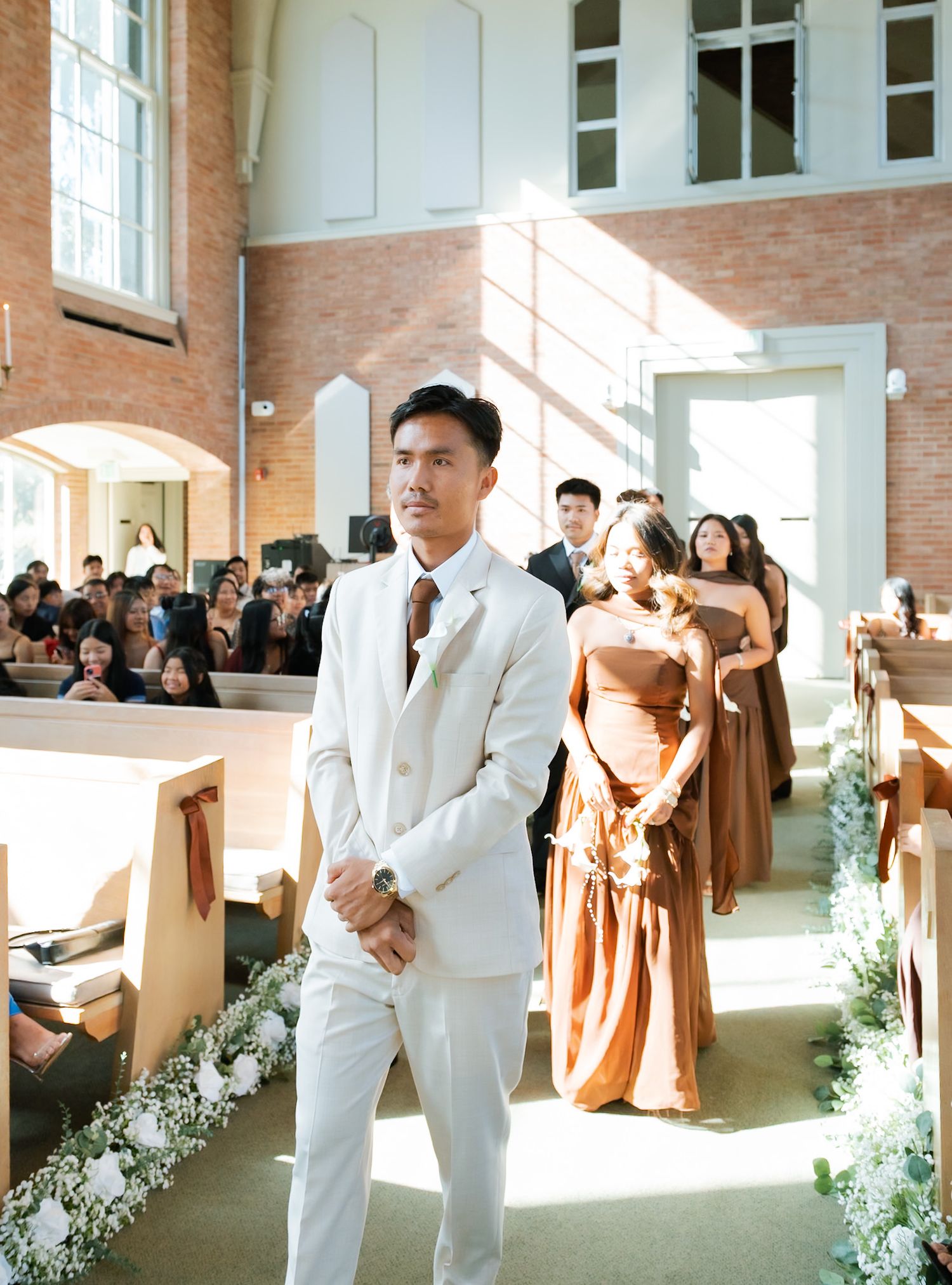 Groom in white suit stands at church altar as bridesmaids in rust dresses walk down the aisle during wedding ceremony.
