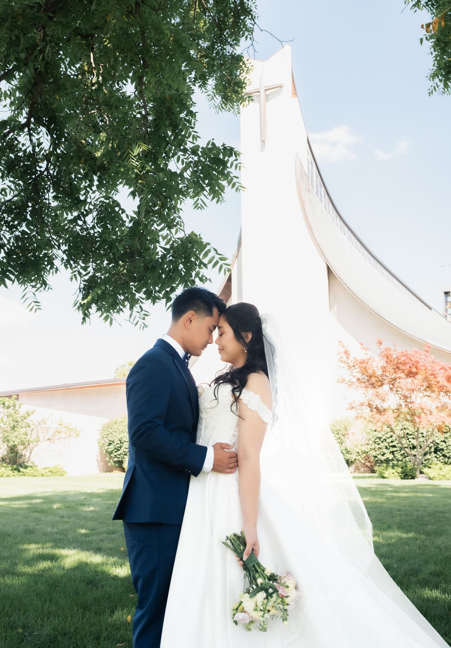 Bride and groom share romantic forehead touch outside church with cross, bride holding white bouquet in elegant wedding attire.