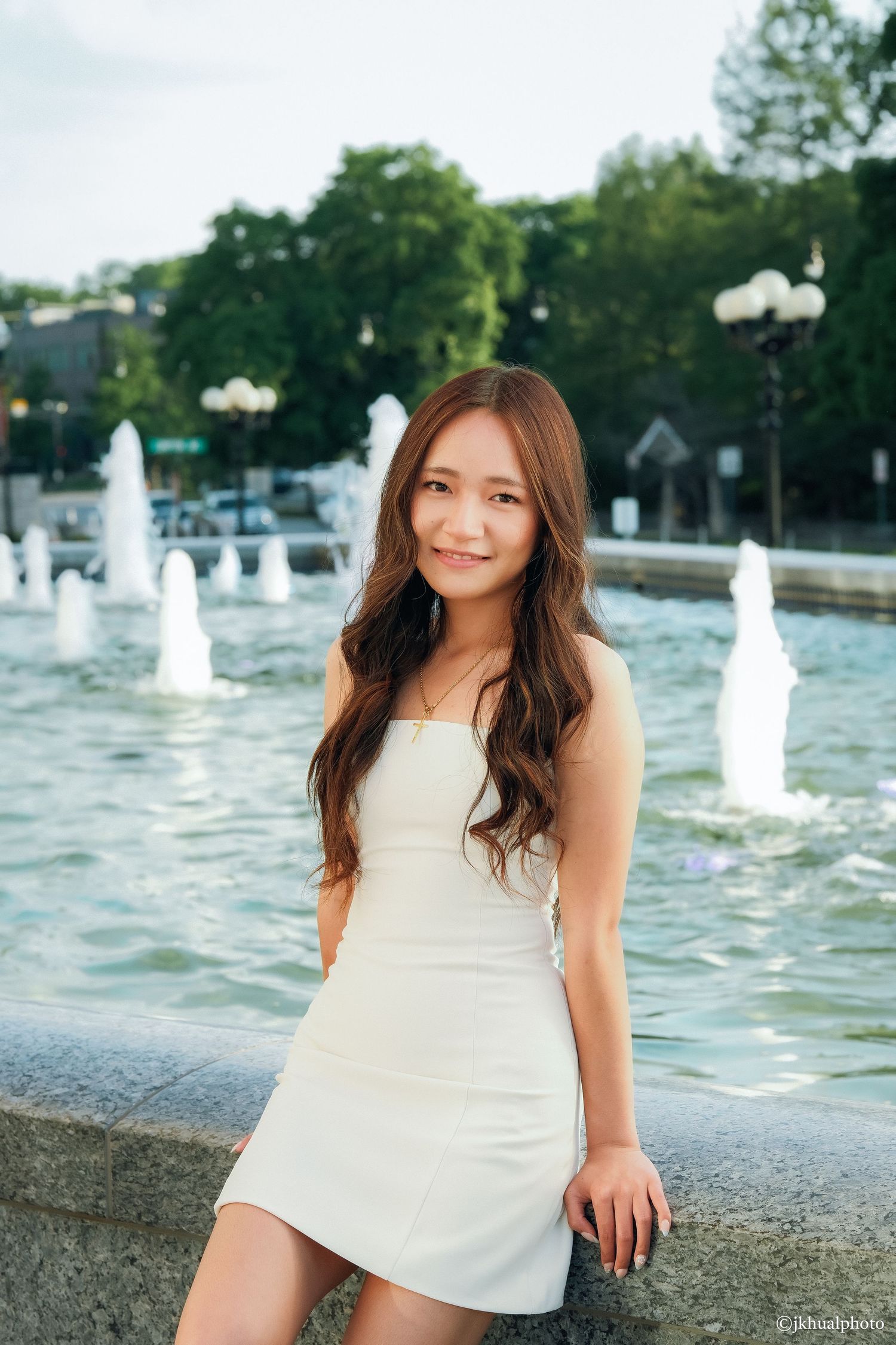 Young woman in white mini dress smiling near fountain with trees in background.