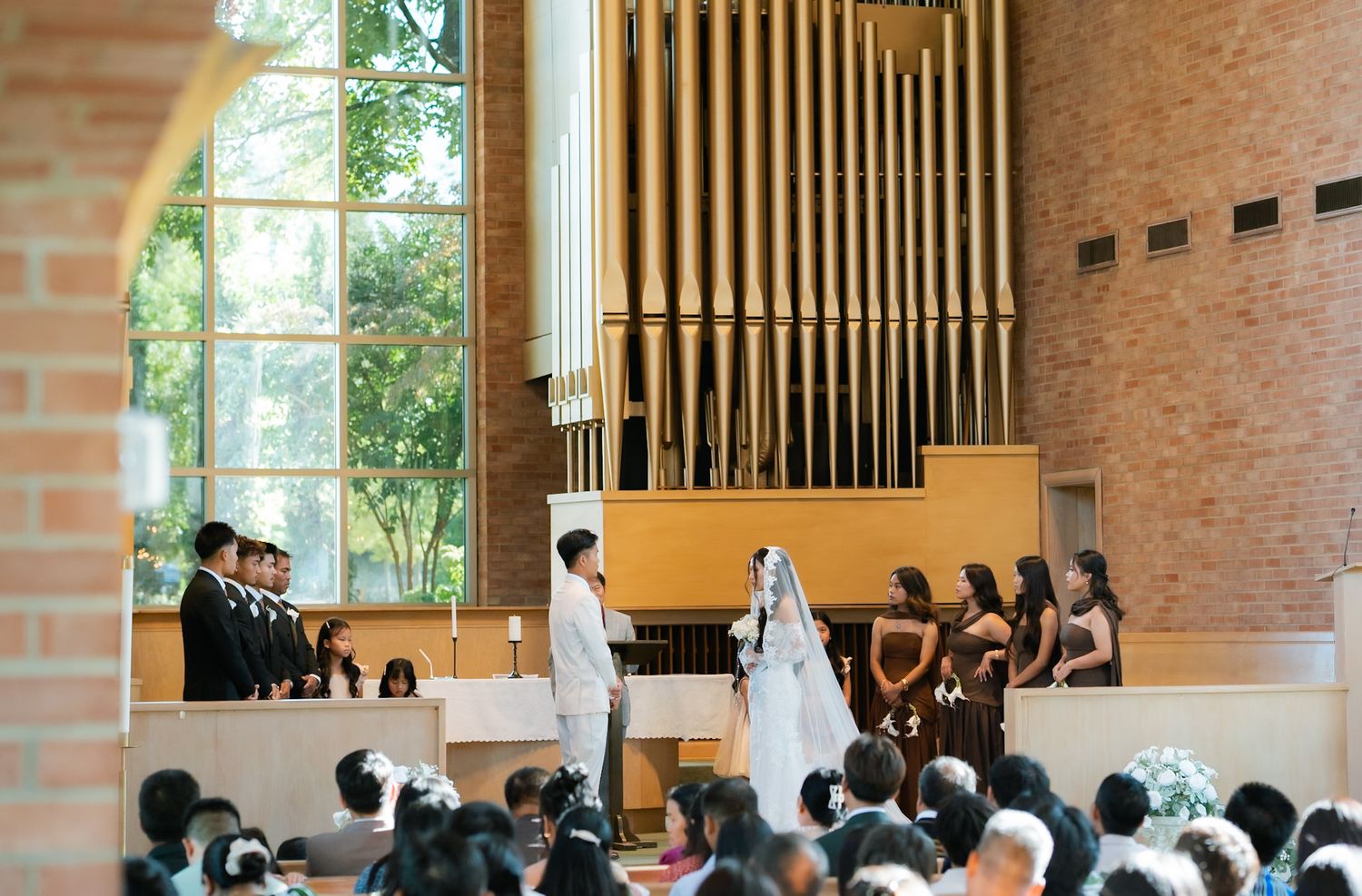 Wedding ceremony in a modern church with organ pipes, bride and groom at altar, bridesmaids, groomsmen, and seated guests.
