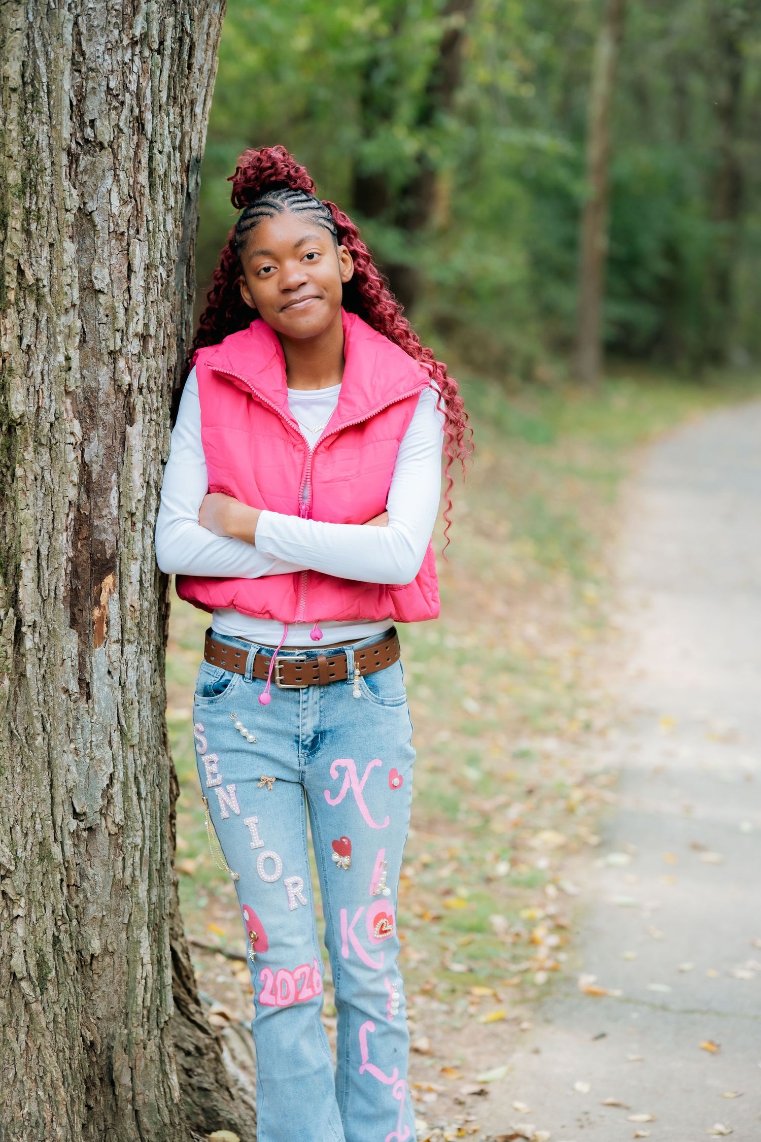 Person in pink vest and decorated jeans smiling while leaning against tree.