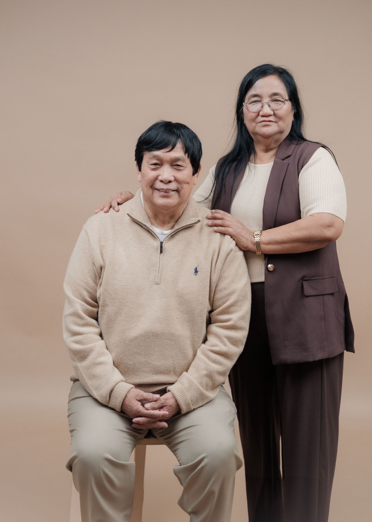 A couple posing together with one seated and one standing in casual attire.