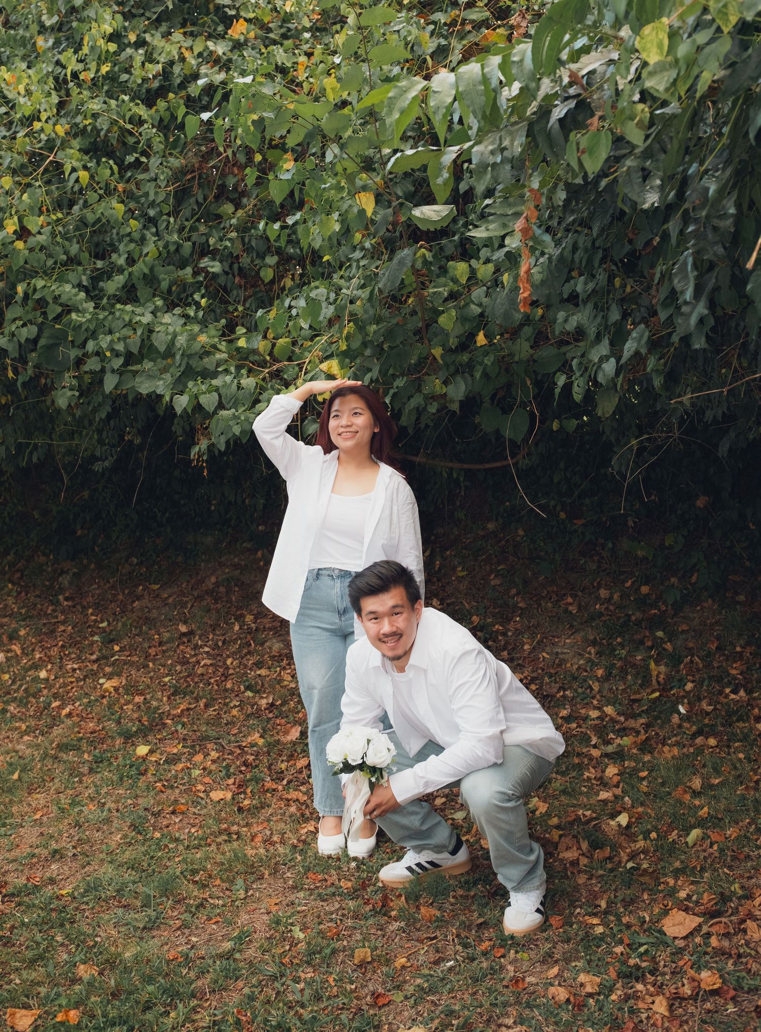 Couple in coordinated white outfits plays in a garden with fallen autumn leaves.