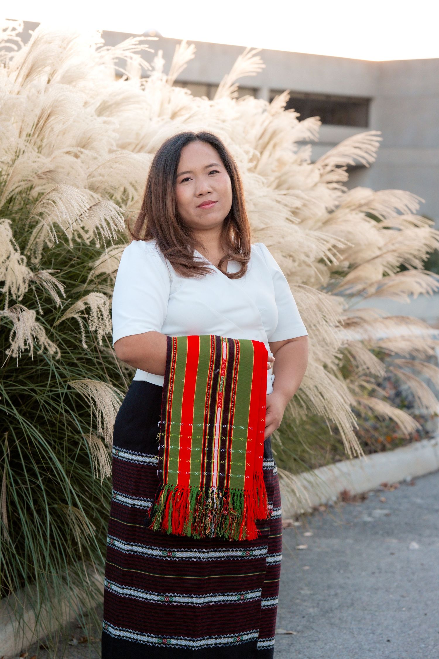 Graduate wearing traditional dress with colorful woven sash poses near ornamental grasses.