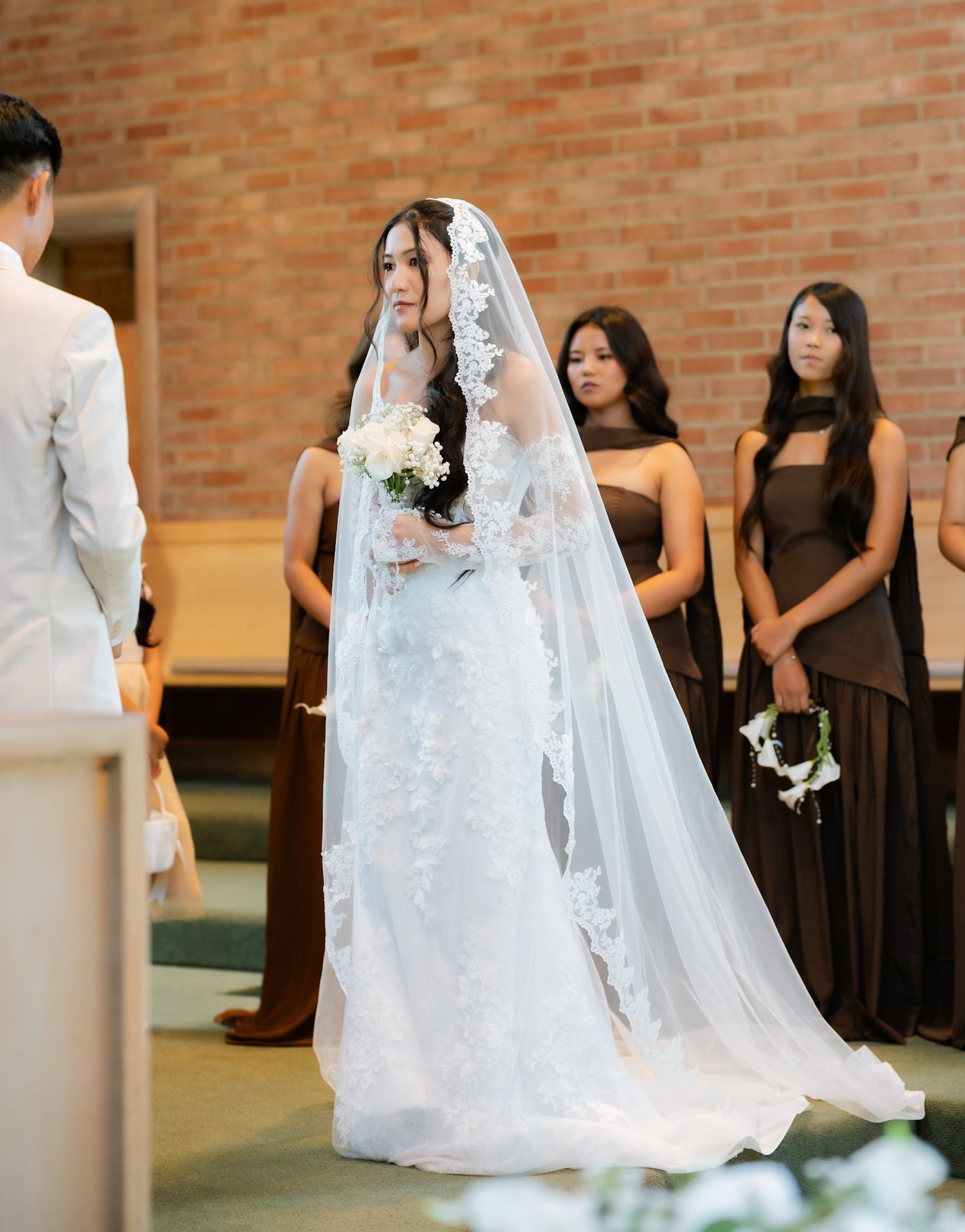 Bride in lace wedding gown and long veil walks down church aisle, bridesmaids in brown dresses standing behind her.